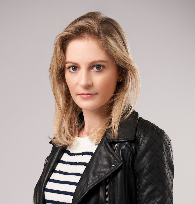 Person with shoulder-length blonde hair wearing a striped shirt and a black leather jacket, standing against a plain background.