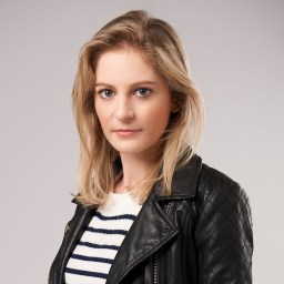 Person with shoulder-length blonde hair wearing a striped shirt and a black leather jacket, standing against a plain background.