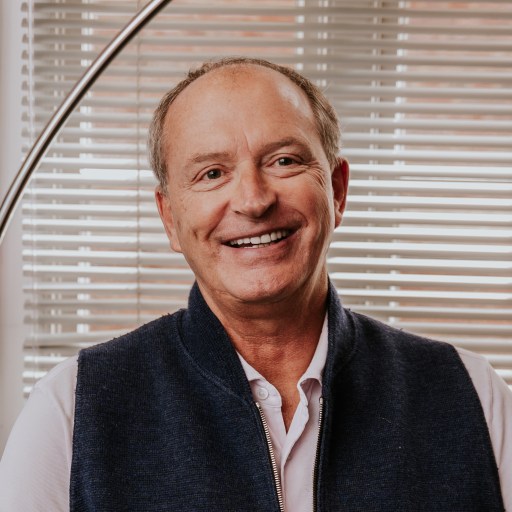 Smiling man in a dark vest and light shirt, exuding leadership, sits indoors in front of window blinds with a curved floor lamp, embodying harmony.