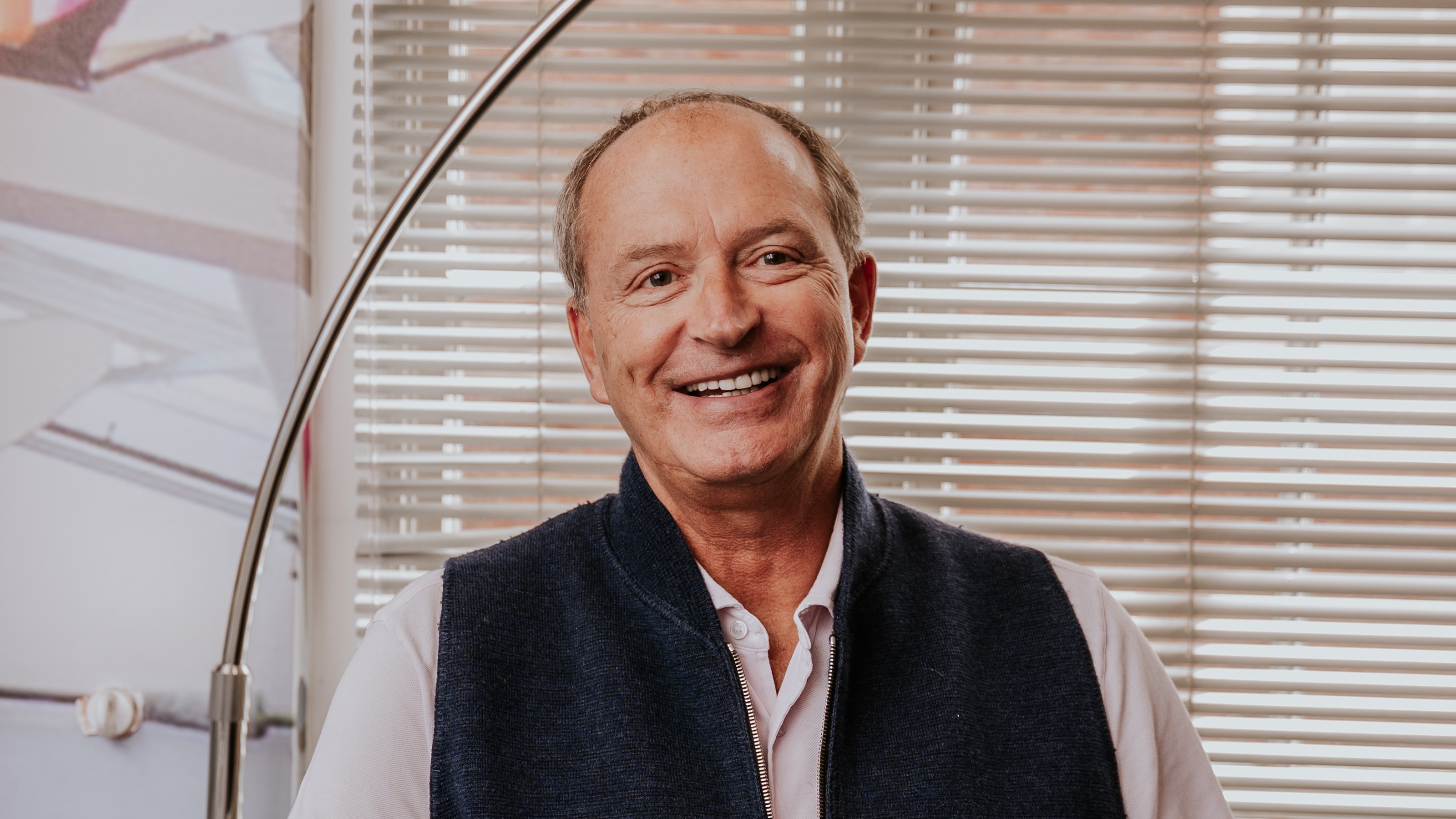 Smiling man in a dark vest and light shirt, exuding leadership, sits indoors in front of window blinds with a curved floor lamp, embodying harmony.
