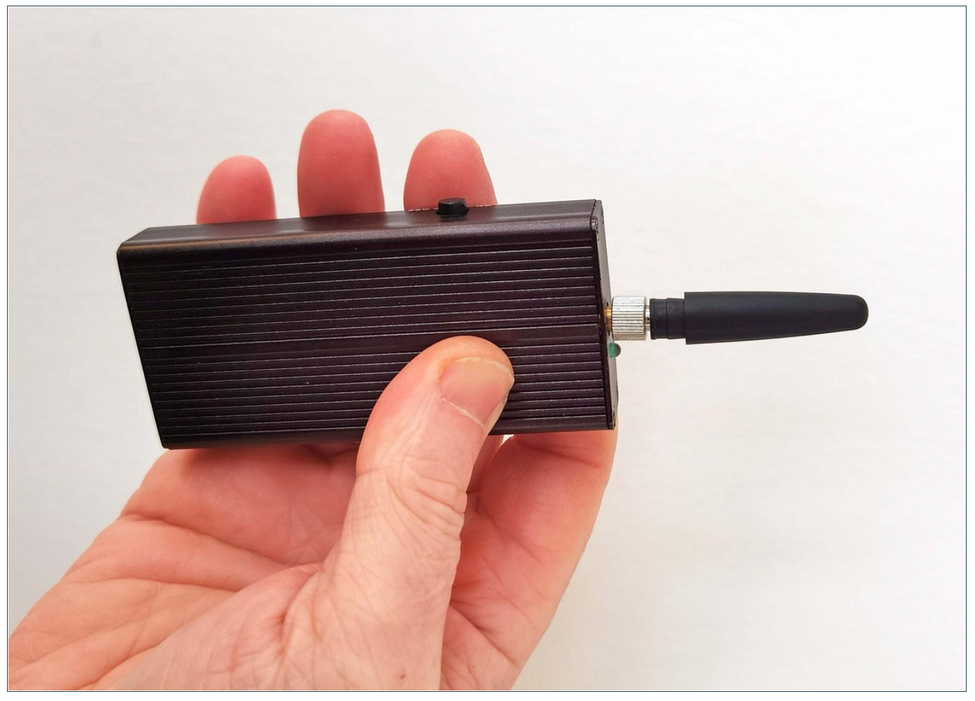 A hand holding a small rectangular black device with a ridged surface and an antenna.