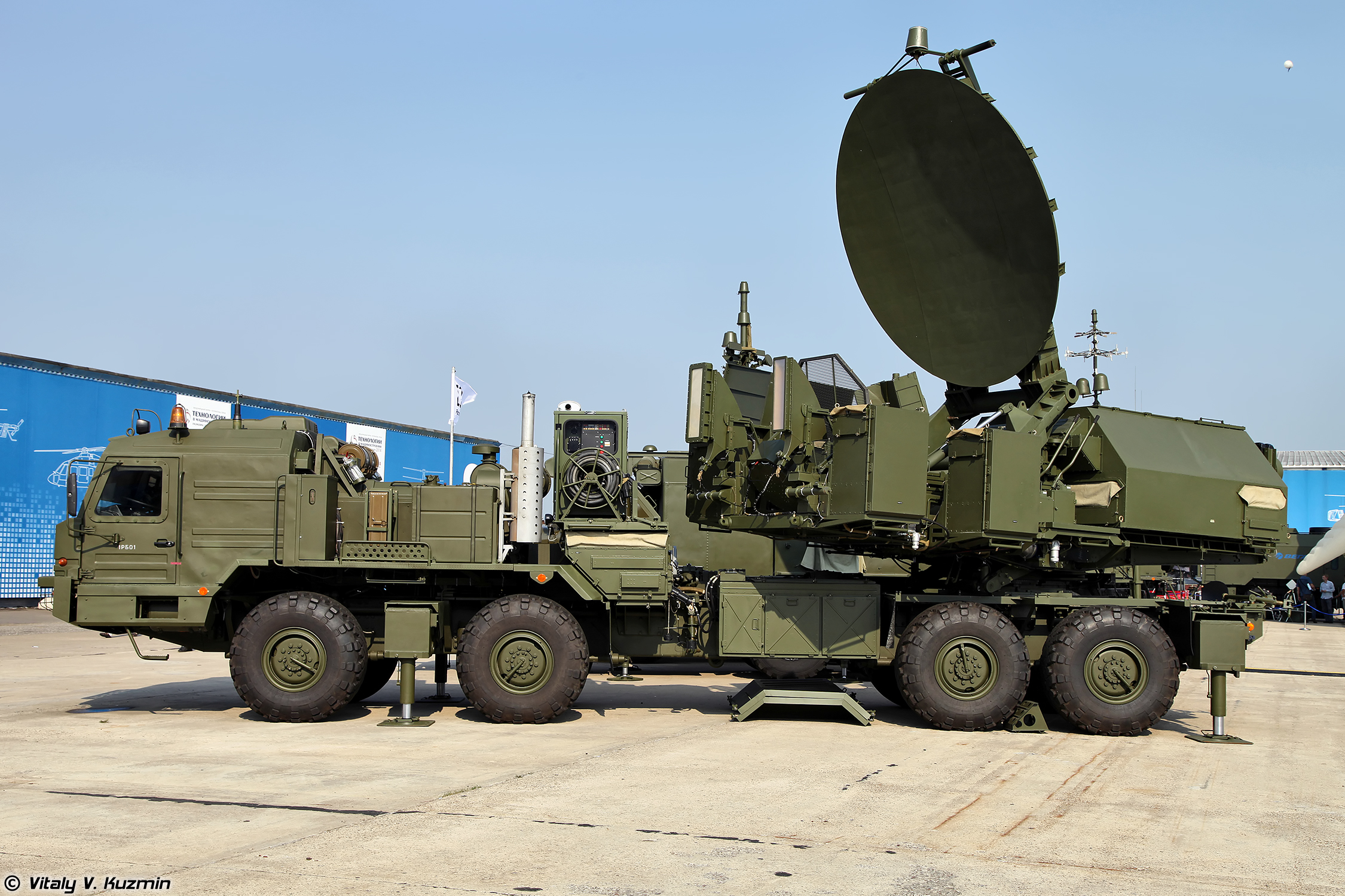 Military radar system mounted on a green, large, multi-wheeled vehicle, displayed outdoors on a concrete surface under clear skies.