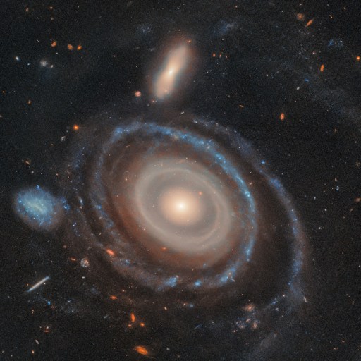 A spiral galaxy with a luminous core, surrounded by swirling arms and smaller galaxies, forms a mesmerizing bullseye ring galaxy, set against a backdrop of stars in space.