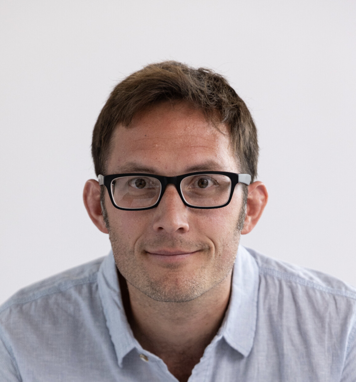 A man with short brown hair and glasses wearing a light blue button-up shirt looks directly at the camera against a plain white background.