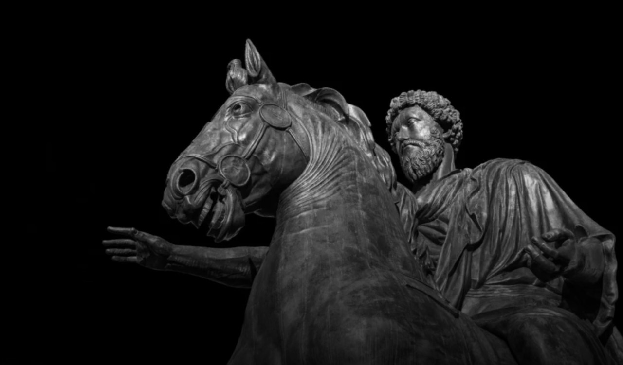 Bronze statue of a bearded man in a robe riding a horse, set against a black background.
