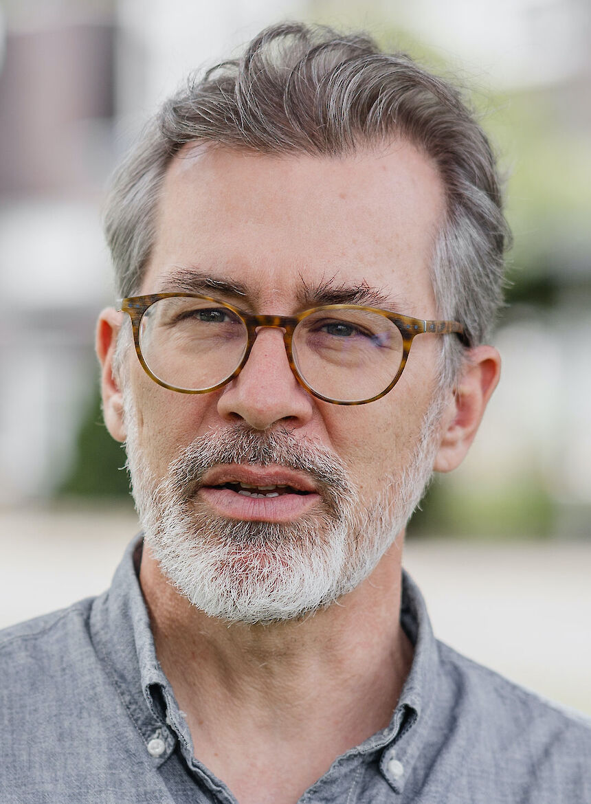 Man with gray hair, beard, and glasses wearing a gray button-up shirt, standing outdoors.