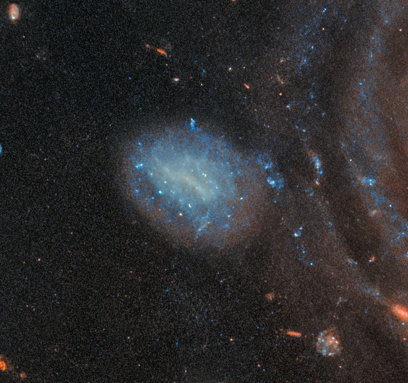 A spiral galaxy with a bright core and diffuse blue outer regions forms a stunning bullseye ring galaxy, surrounded by stars and distant celestial objects against the dark void of space.