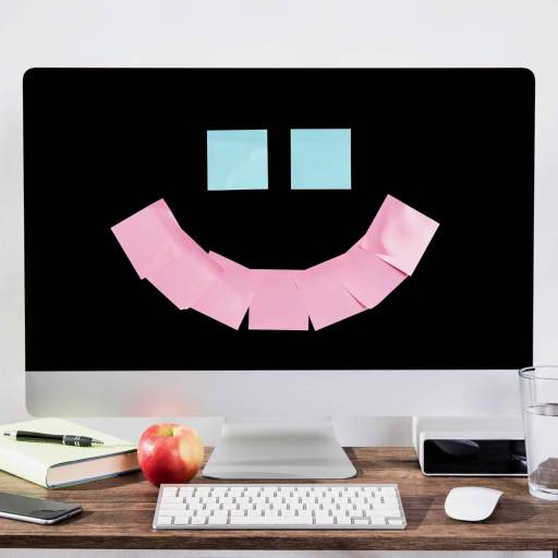 A computer screen adorned with a smiley face made of sticky notes exudes workplace happiness, sitting on a desk alongside a keyboard, mouse, phone, notebook, apple, glass of water, and small plant.