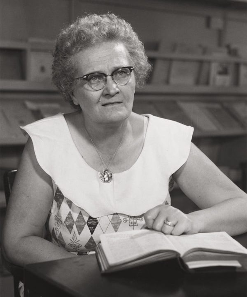 A woman with glasses and curly hair, reminiscent of Cecilia Payne's intellectual charm, sits at a table wearing a patterned dress. She holds an open book in front of a bookshelf, seemingly lost in thought among 100 stories.