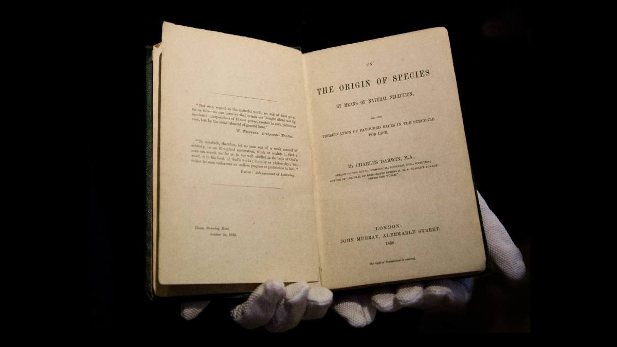 Gloved hands hold an open book titled "The Origin of Species by Means of Natural Selection" by Charles Darwin, a seminal work exploring the origin of life, published in London by John Murray, 1859.
