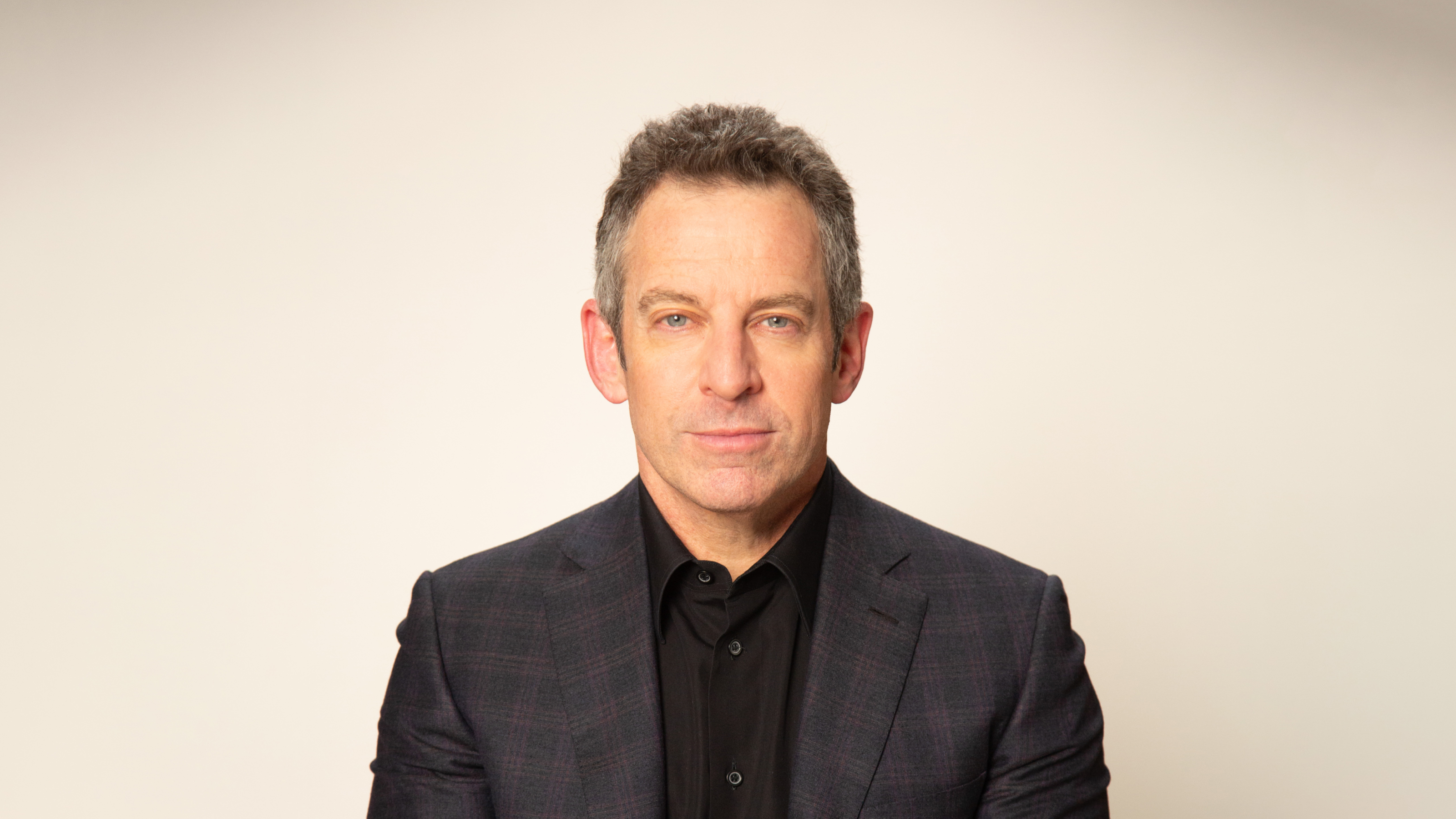 A person in a black shirt and dark blazer poses against a plain background.