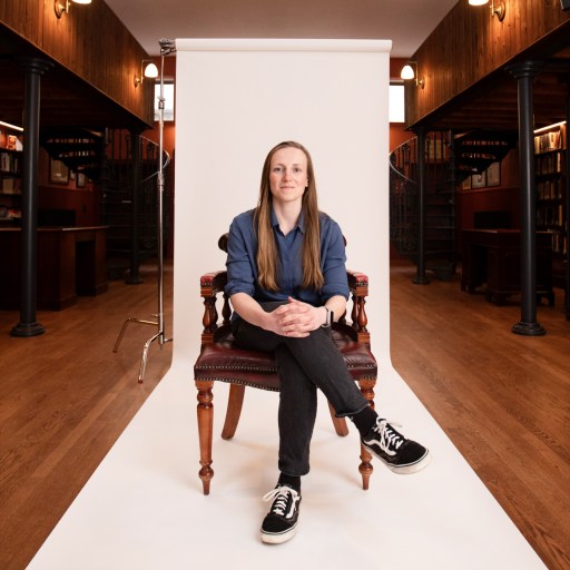 A person sits on a chair in a library setting, with bookshelves on either side and a white backdrop behind them.