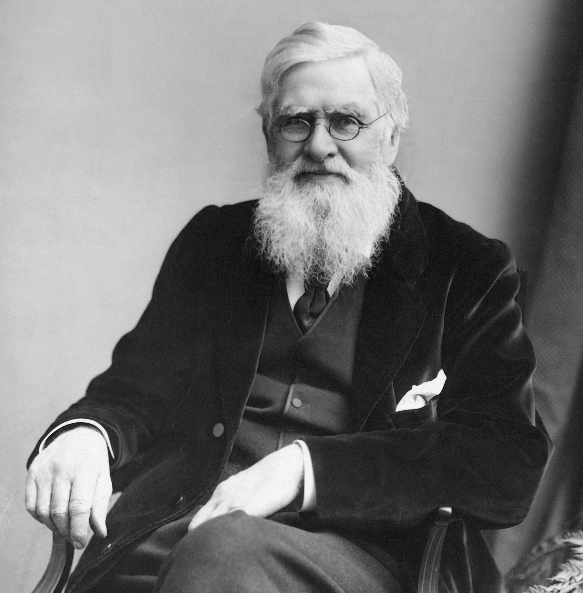 Elderly man with a long white beard, seated in a three-piece suit and glasses, looks at the camera.