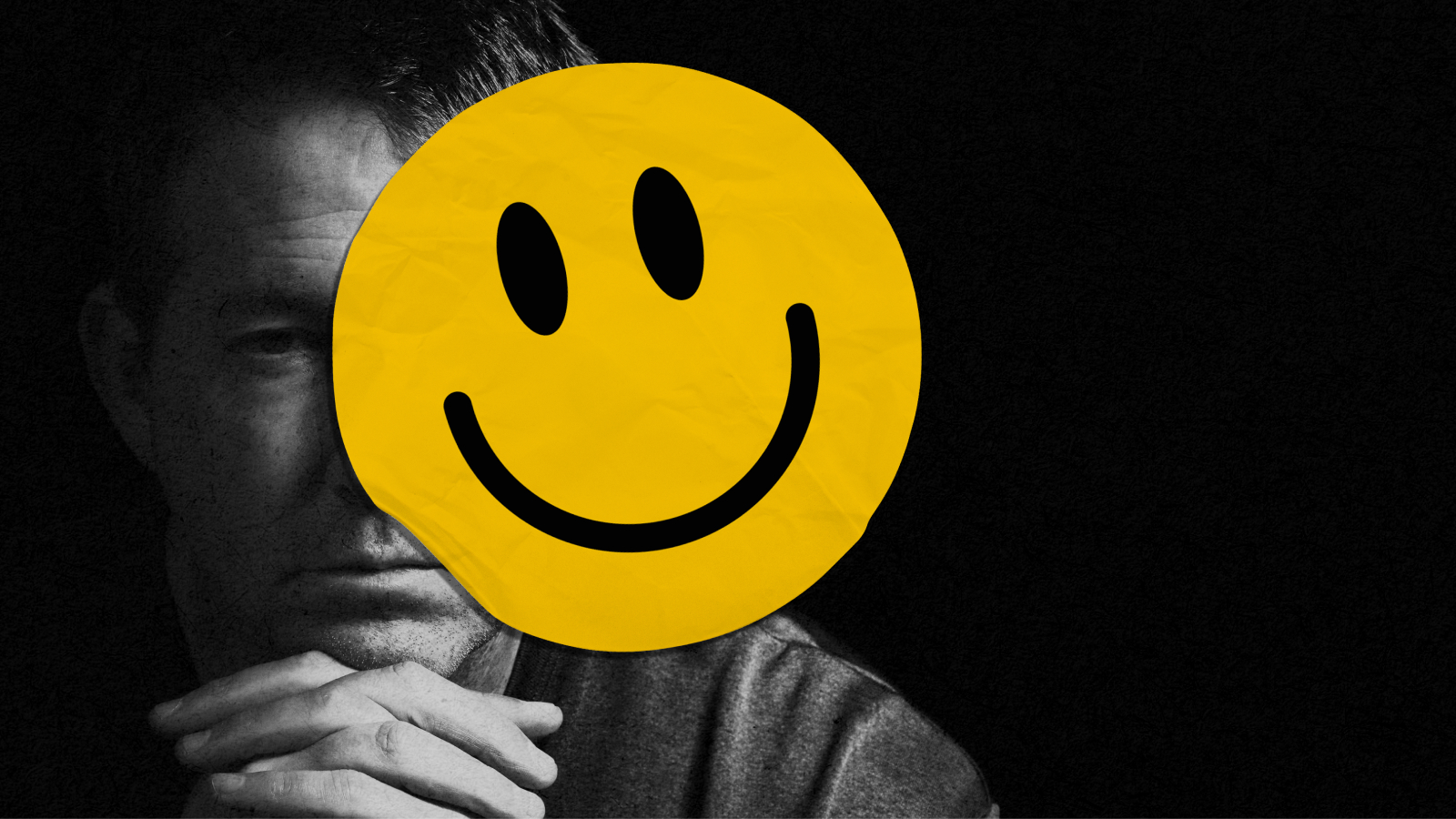 A person sits in darkness, frowning, partially obscured by a large yellow smiley face sticker covering part of their face.