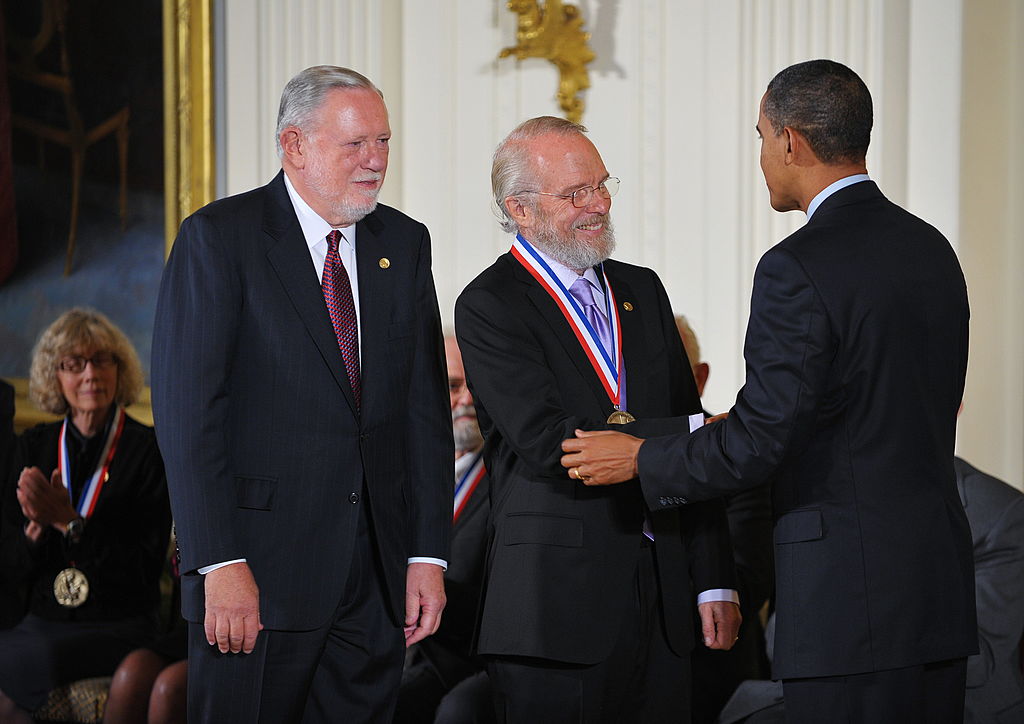 A man in a suit shakes hands with another man wearing a medal, while a third man stands beside them. An audience member claps in the background.