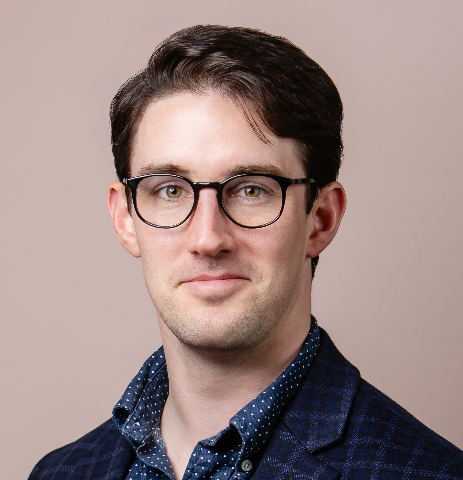A person with short dark hair and glasses, wearing a dark blazer over a polka dot shirt, in front of a beige background.