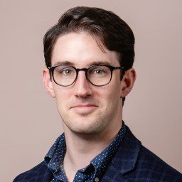 A person with short dark hair and glasses, wearing a dark blazer over a polka dot shirt, in front of a beige background.