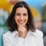 A person with shoulder-length dark hair, wearing hoop earrings and a white shirt, smiles while resting their chin on their hand. The background is blurred and colorful.