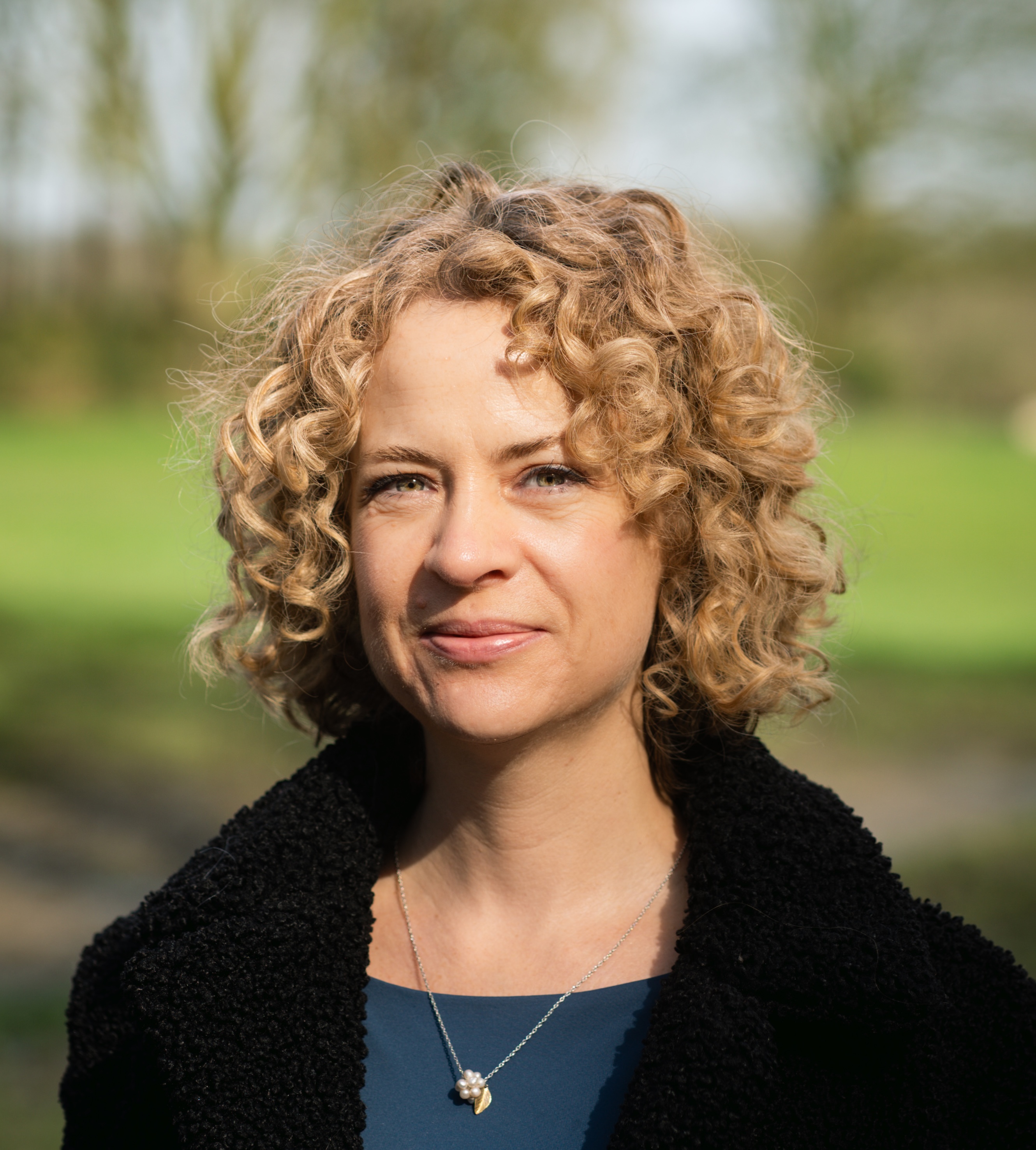 Woman with curly hair wearing a dark jacket and necklace, standing outdoors with trees and grass in the background.