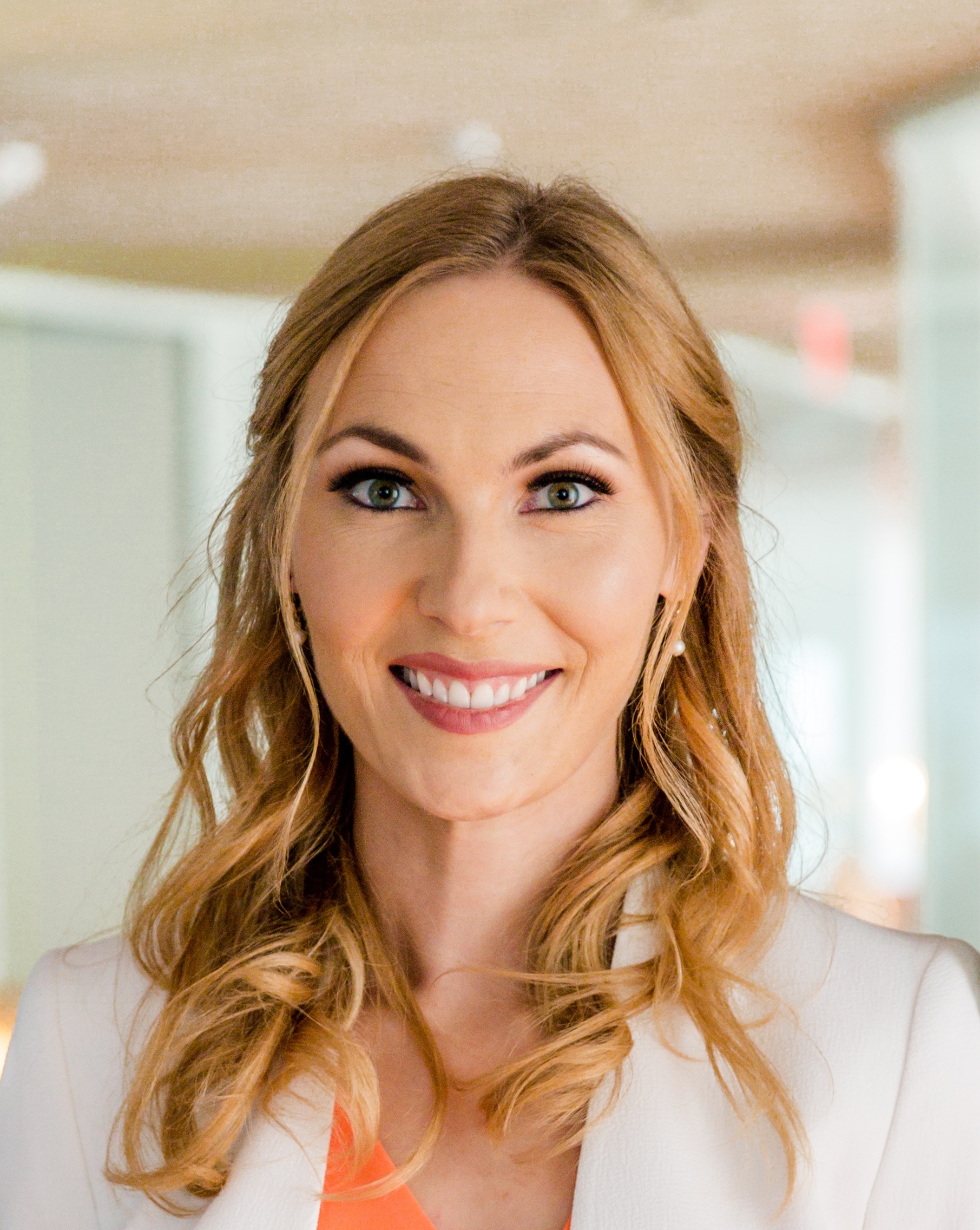 A woman with long blonde hair smiles at the camera, wearing a white blazer and coral top, with blurred office-like background.