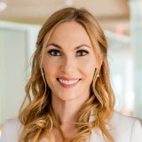 A woman with long blonde hair smiles at the camera, wearing a white blazer and coral top, with blurred office-like background.