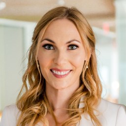 A woman with long blonde hair smiles at the camera, wearing a white blazer and coral top, with blurred office-like background.
