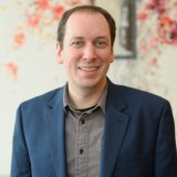 A person in a blue blazer and gray shirt stands smiling in front of a colorful abstract background.