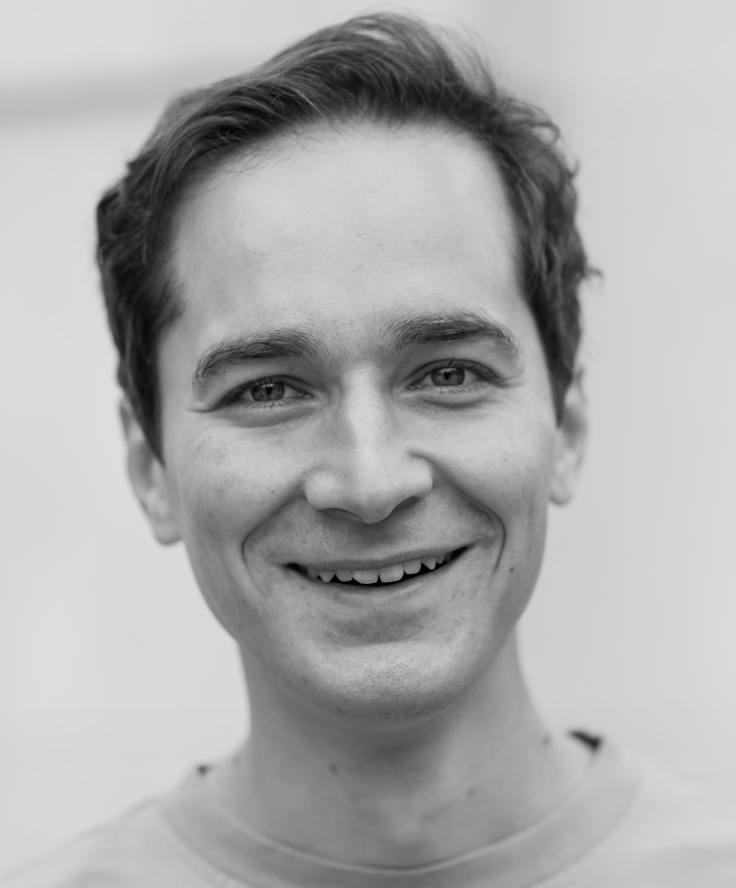 A black and white portrait of a person smiling, wearing a light-colored shirt, with a neutral background.