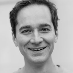 A black and white portrait of a person smiling, wearing a light-colored shirt, with a neutral background.