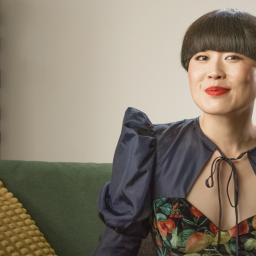 A woman with a bowl haircut and red lipstick sits on a sofa wearing a dark, floral dress. A textured yellow pillow and patterned wall are visible in the background.