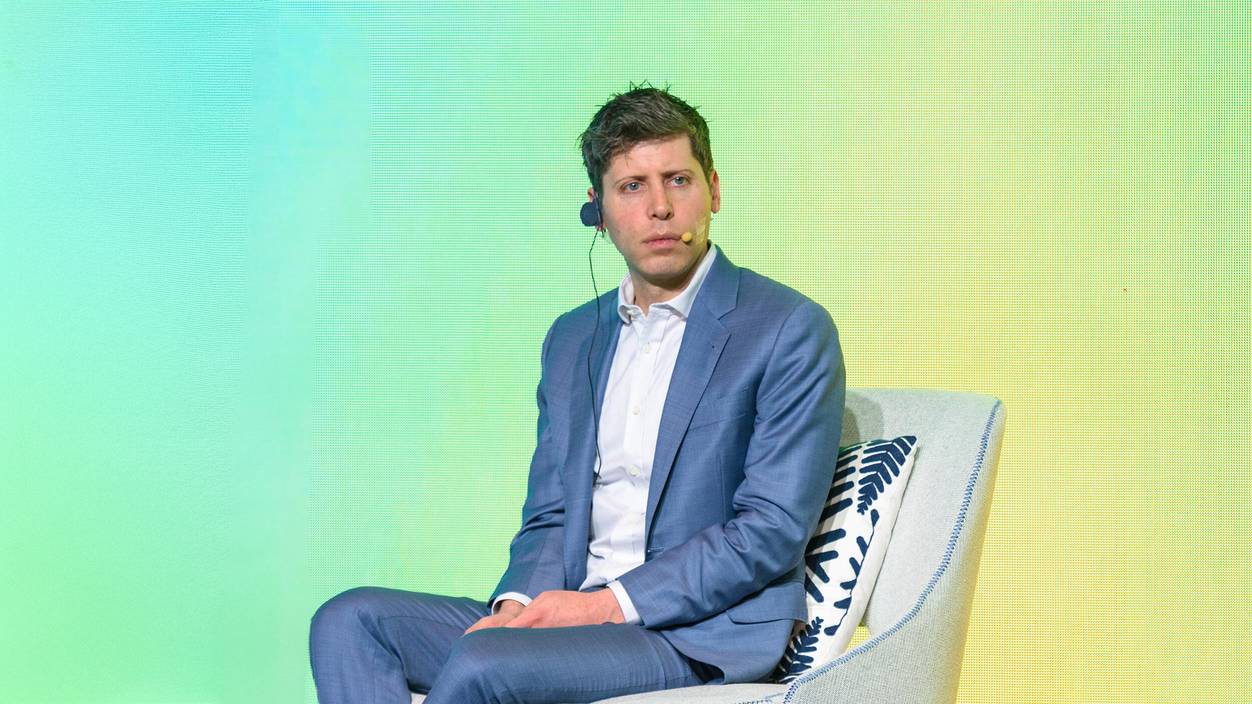 A man in a suit sits on a stage chair with a headset, set against a vibrant green background, ready to discuss the latest advancements at OpenAI.