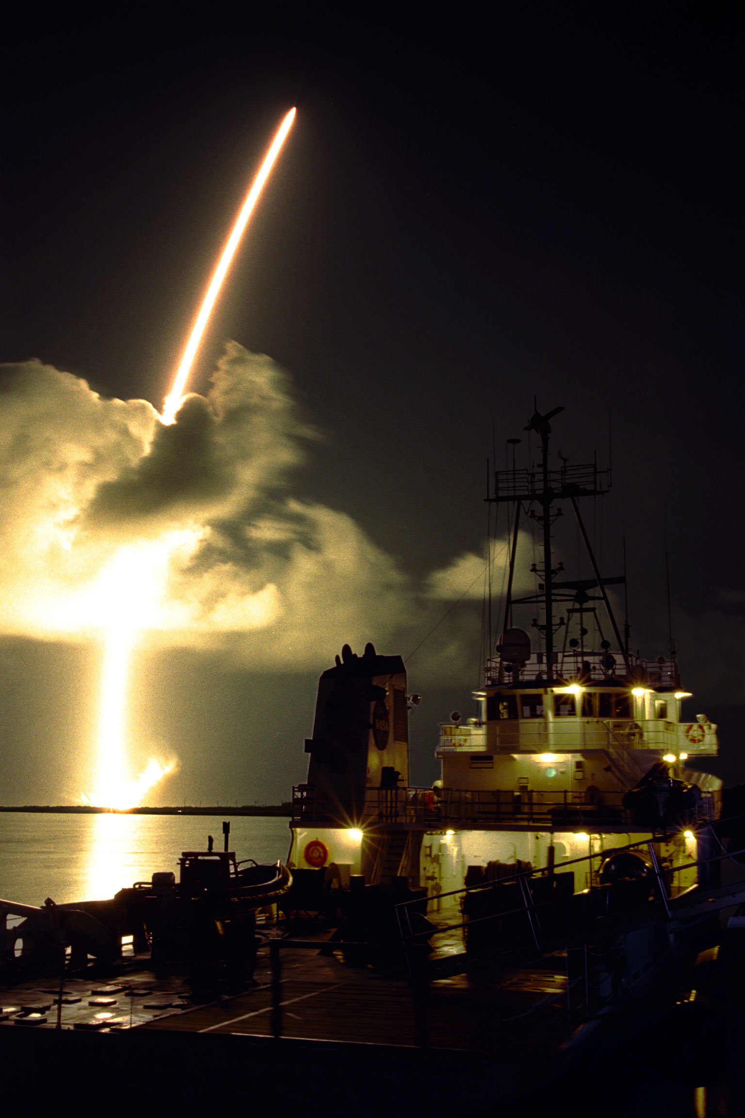 A rocket launches into the night sky over the ocean, leaving a bright trail, showcasing the fruits of fundamental research. A ship is illuminated in the foreground.