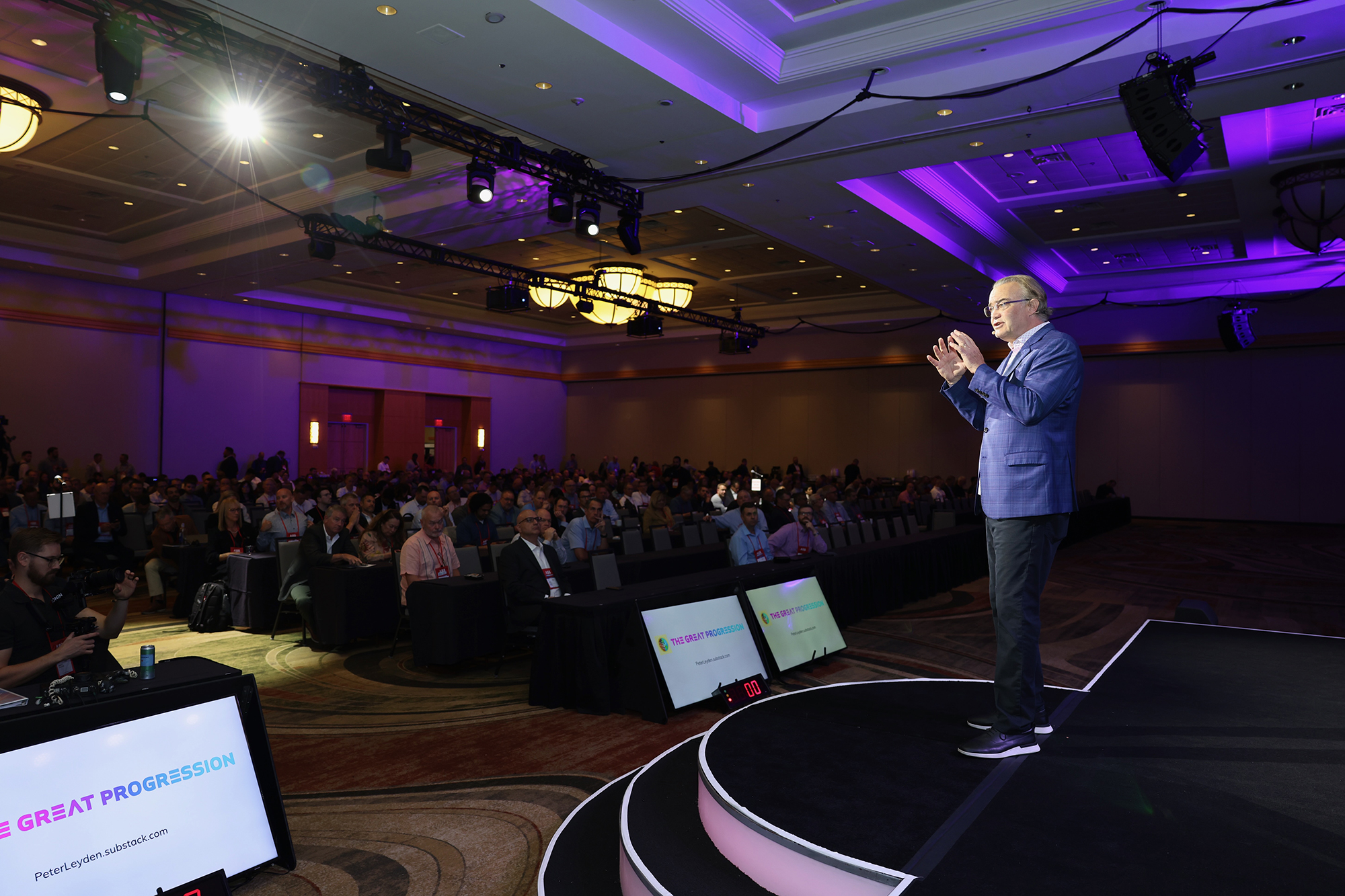 Man speaking on stage at a conference, addressing a large seated audience in a venue with purple ceiling lights. Multiple screens display event branding.