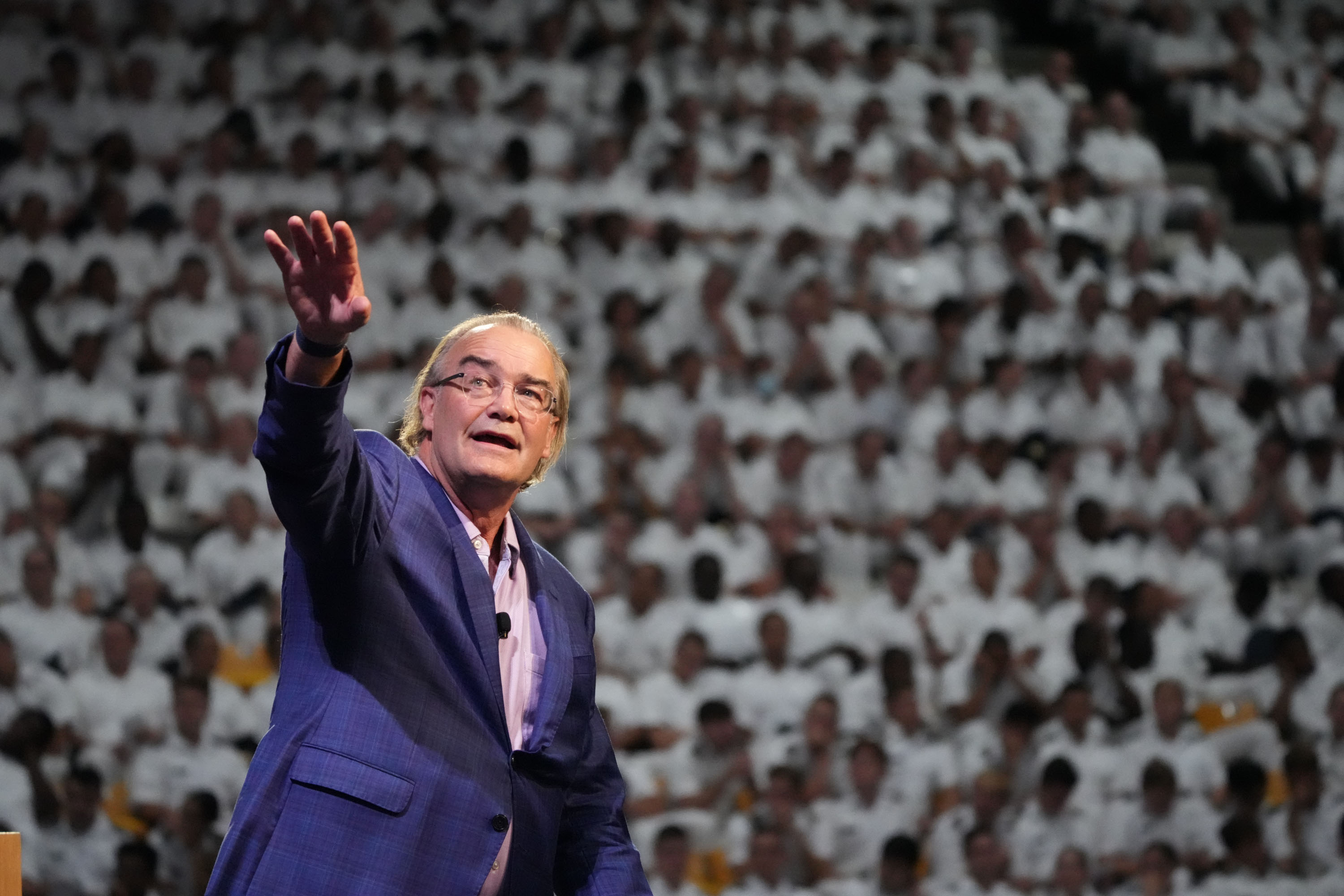 A man in a suit speaks on stage, gesturing with his hand, in front of a large audience wearing white uniforms.
