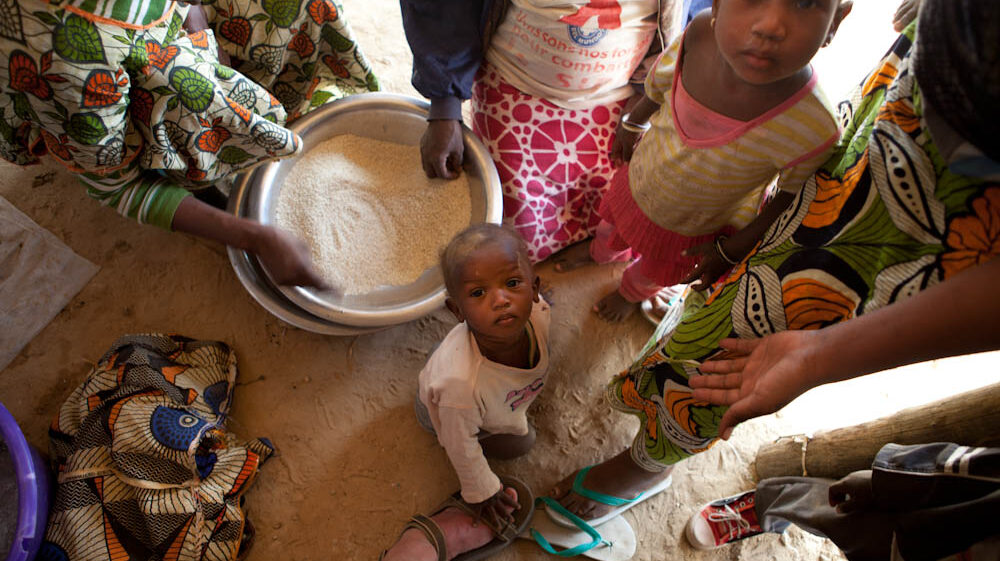 Children and adults gather around a large bowl of rice placed on the ground, like researchers investing their time in fundamental bonds. A child kneels nearby, looking up. The scene unfolds indoors on a dirt floor.