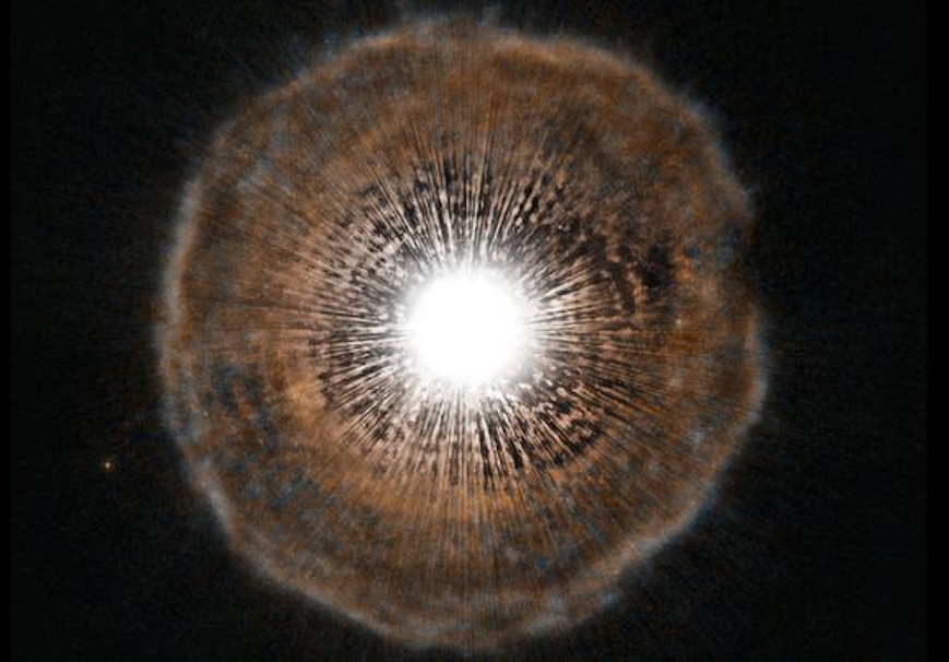 Image of a bright white star at the center, resembling a sun in the final stages as it transforms into a planetary nebula. A diffuse halo of brown and orange colors envelops it against a dark backdrop, illustrating the beauty that remains as a star dies.