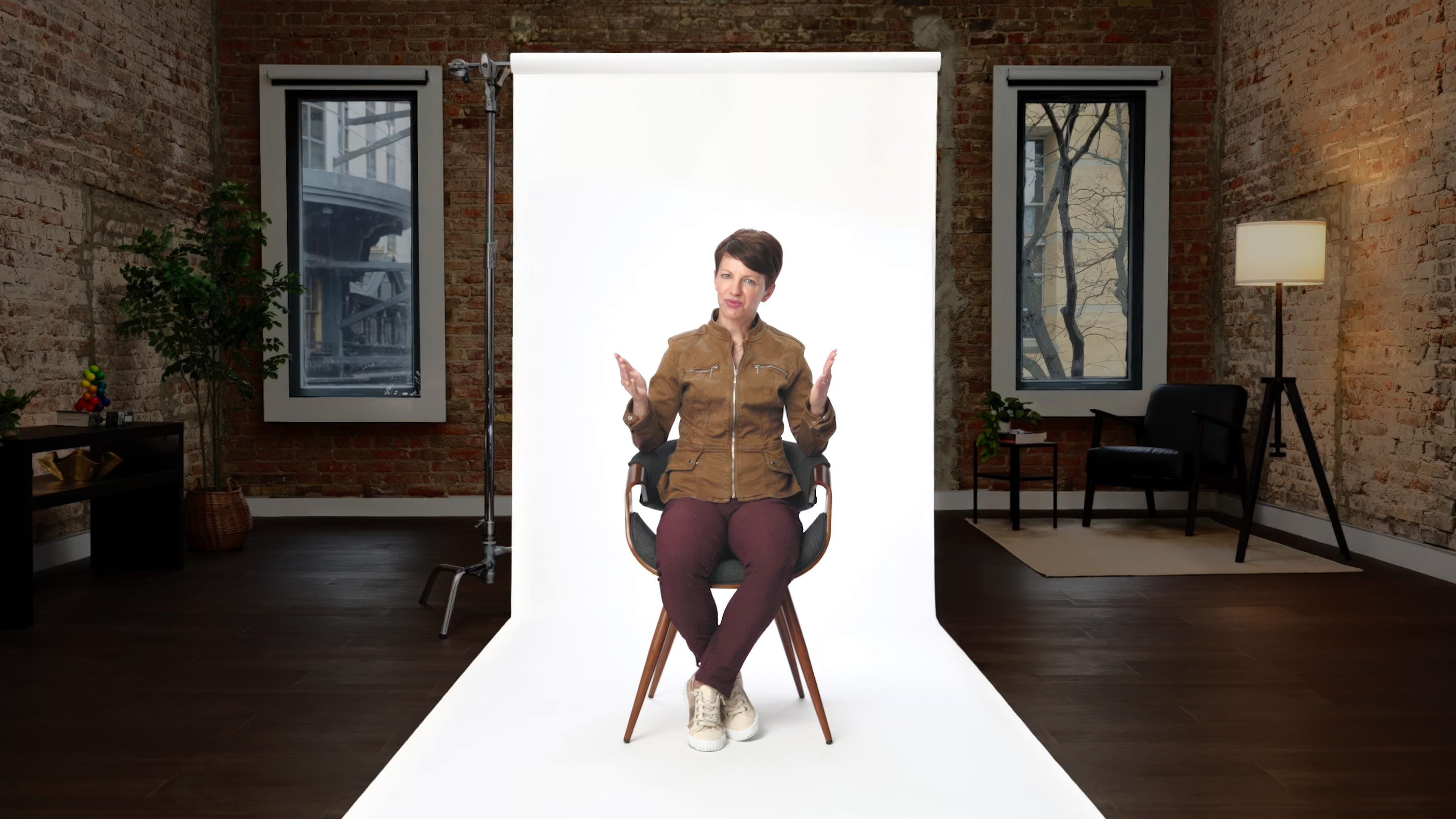 A person sits on a chair in front of a white backdrop in a loft-style room with brick walls, dark flooring, and two windows.