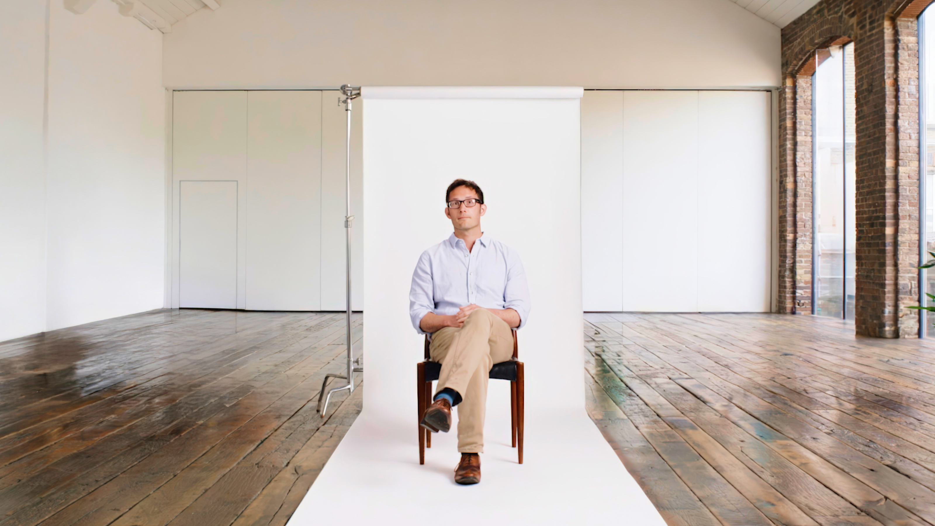 A person sitting on a chair with legs crossed in front of a white backdrop in a spacious room with wooden floors and large windows.