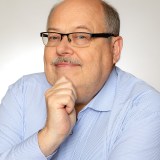 A man wearing glasses and a light blue shirt with vertical stripes, resting his chin on his hand, poses against a plain background.