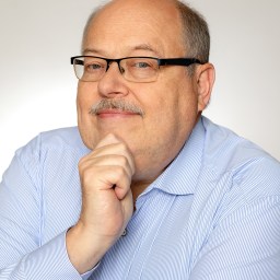 A man wearing glasses and a light blue shirt with vertical stripes, resting his chin on his hand, poses against a plain background.