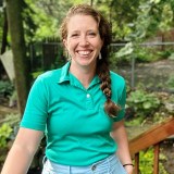 Person with long braided hair smiles at the camera wearing a turquoise polo shirt and light pants, standing outdoors with trees and plants in the background.