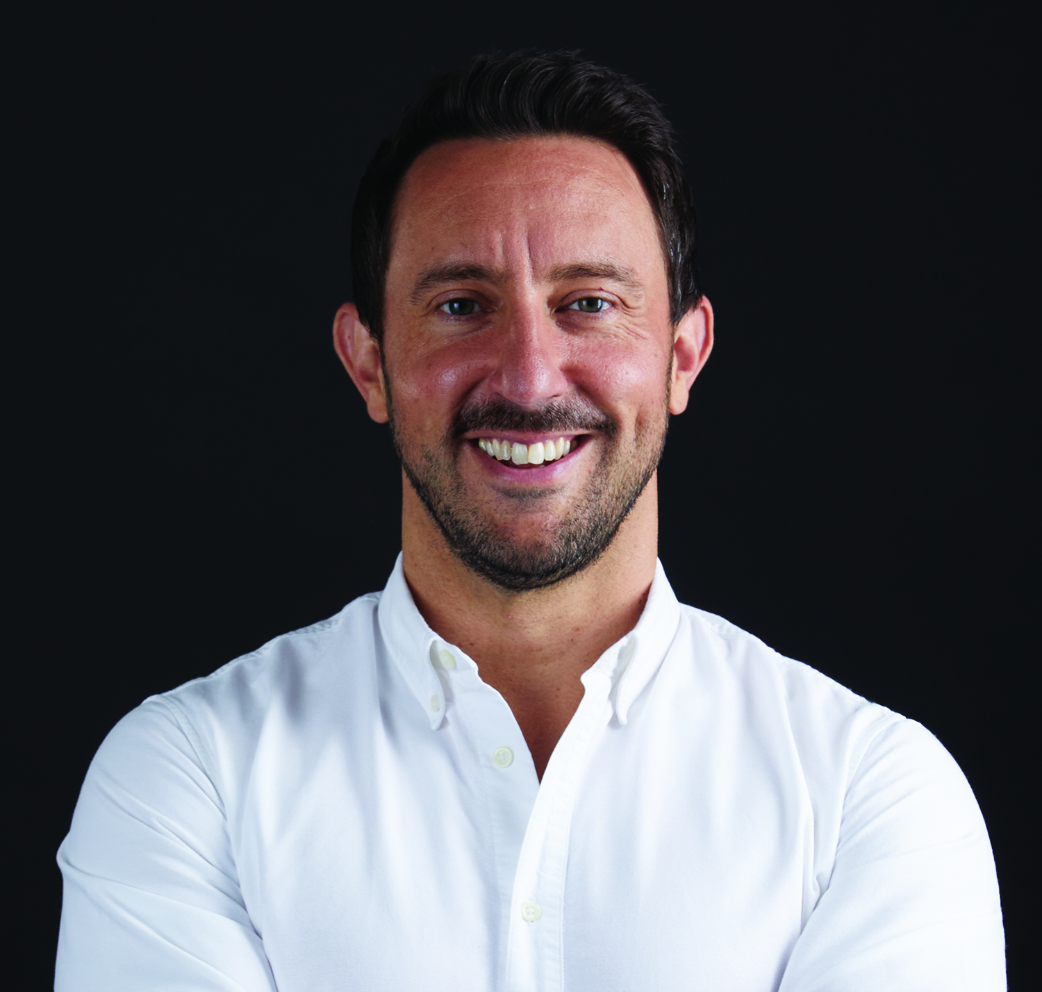 A man with short dark hair and facial hair smiles, wearing a white button-up shirt, against a black background.