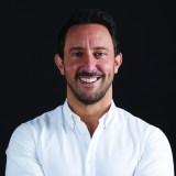 A man with short dark hair and facial hair smiles, wearing a white button-up shirt, against a black background.