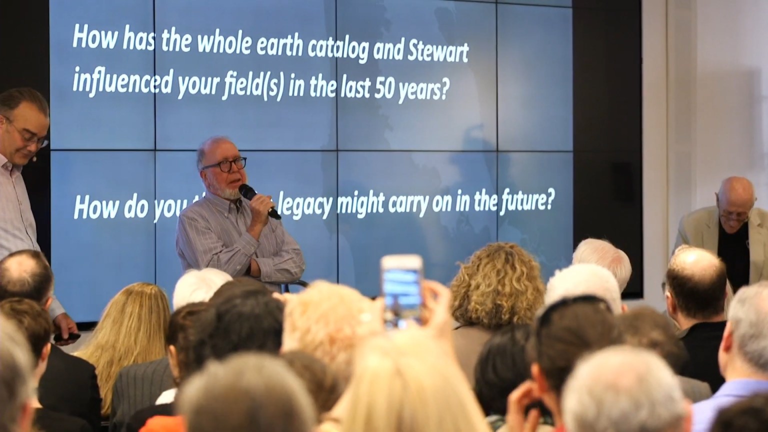 A group of people in a room attends a talk. A speaker holds a microphone in front of a screen displaying questions about the influence and future legacy of a catalog and Stewart.