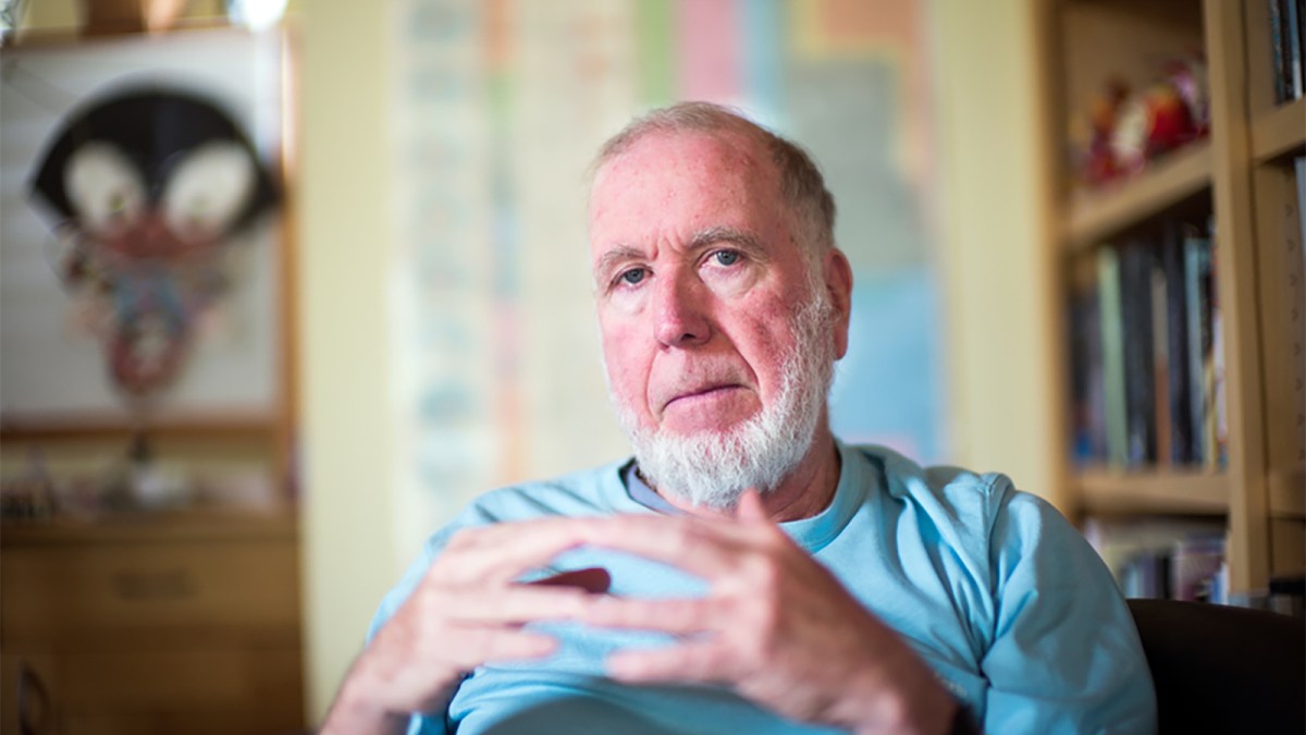 An older man with a white beard sits in a room, wearing a light blue shirt, hands clasped together. Bookshelves and artwork are visible in the background.