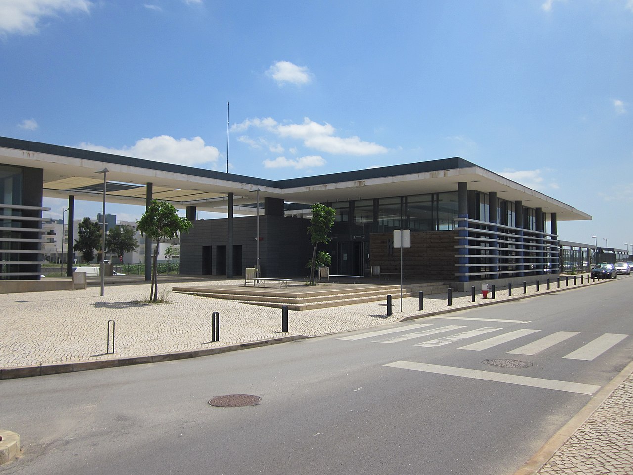 Modern, low-rise building with large windows and flat roof, featuring a paved walkway, young trees, and a pedestrian crosswalk under a clear blue sky.