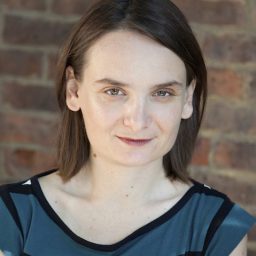 A woman with straight brown hair and light skin wearing a blue and black striped top stands in front of a brick wall, looking directly at the camera.