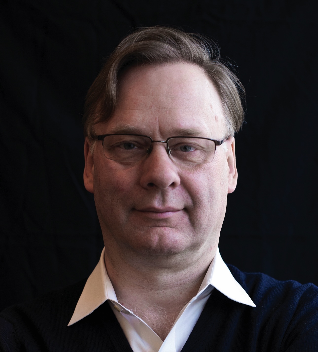 A person wearing glasses and a dark sweater over a white shirt against a dark background.