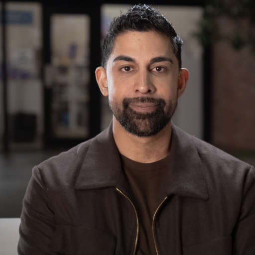 A person with short dark hair and a beard sits indoors wearing a brown jacket and dark shirt. The background features blurred indoor elements and a tree.