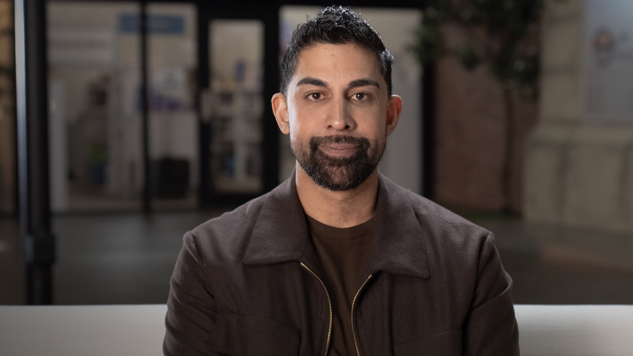A person with short dark hair and a beard sits indoors wearing a brown jacket and dark shirt. The background features blurred indoor elements and a tree.